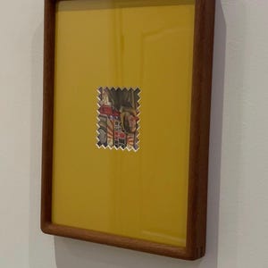 May include: A framed artwork featuring a yellow background with a small, postage stamp-like image in the center. The frame is made of dark wood. The image on the stamp appears to be a portrait.