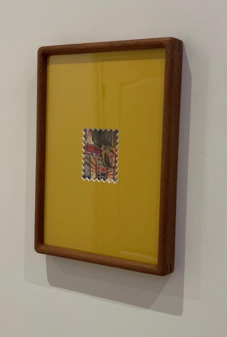 May include: A framed artwork featuring a yellow background with a small, postage stamp-like image in the center. The frame is made of dark wood. The image on the stamp appears to be a portrait.