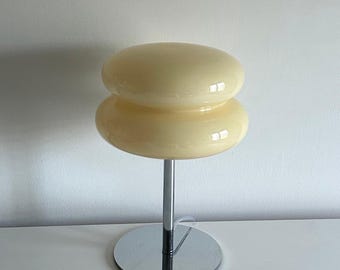 May include: A retro-style table lamp with a stacked, rounded, cream-colored shade. The shade sits atop a chrome stem and a circular base. The lamp's design is reminiscent of mid-century modern aesthetics, offering a warm, ambient light source.