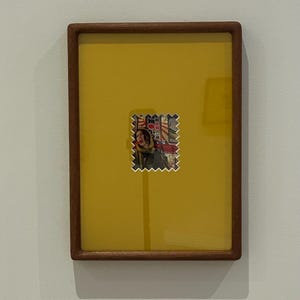 May include: A framed artwork featuring a yellow background with a small, colorful image resembling a postage stamp. The stamp-like image depicts a person and Japanese text. The frame is made of dark wood.