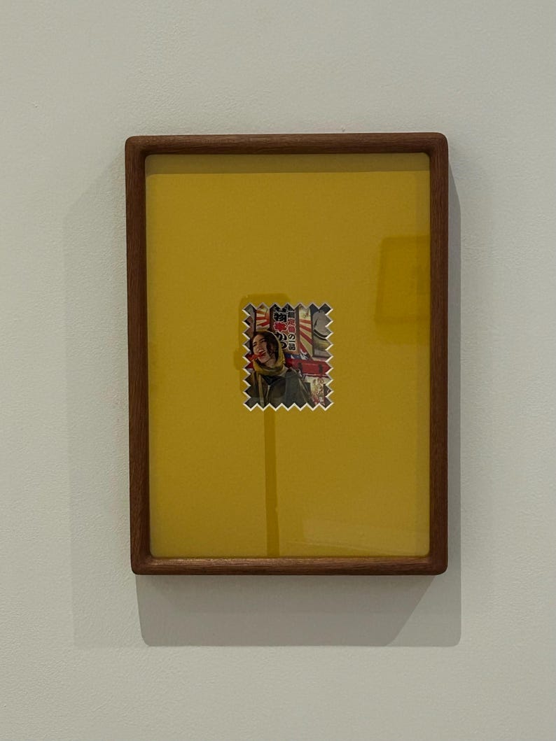 May include: A framed artwork featuring a yellow background with a small, colorful image resembling a postage stamp. The stamp-like image depicts a person and Japanese text. The frame is made of dark wood.
