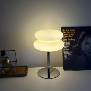 May include: A modern, white glass table lamp with a stacked, disc-shaped shade and a chrome base. The lamp is illuminated, casting a warm glow. A vinyl record is visible in the background.