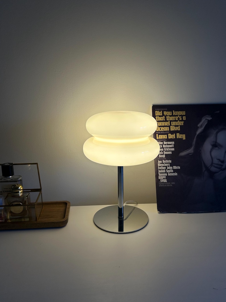 May include: A modern, white glass table lamp with a stacked, disc-shaped shade and a chrome base. The lamp is illuminated, casting a warm glow. A vinyl record is visible in the background.