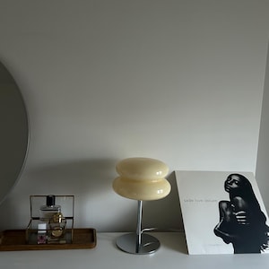 May include: A cream-colored, double-layered mushroom-shaped table lamp with a chrome base. Beside the lamp is a framed perfume set on a wooden tray and a Sade "Love Deluxe" album cover.