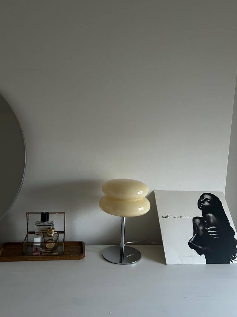 May include: A cream-colored, double-layered mushroom-shaped table lamp with a chrome base. Beside the lamp is a framed perfume set on a wooden tray and a Sade "Love Deluxe" album cover.
