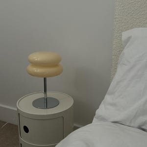 May include: A cream-colored bedside lamp with a stacked, rounded shade sits atop a chrome stem. The lamp is placed on a matching cream-colored nightstand with two drawers. The room has a minimalist aesthetic with a white wall and bed linens.