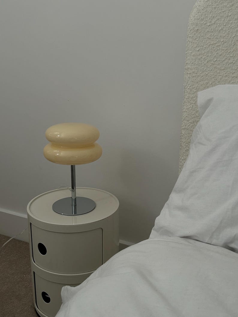 May include: A cream-colored bedside lamp with a stacked, rounded shade sits atop a chrome stem. The lamp is placed on a matching cream-colored nightstand with two drawers. The room has a minimalist aesthetic with a white wall and bed linens.