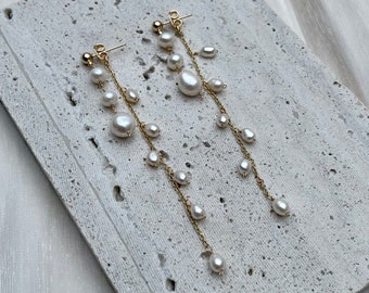 May include: A pair of gold-colored, dangling earrings with multiple white pearls. The earrings are displayed on a light gray, textured surface against a white, silky background. The pearls vary in size and are strung along delicate chains.