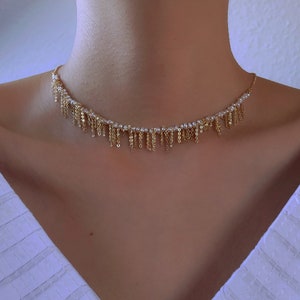 May include: A gold chain necklace with small white pearls and dangling gold chains. The necklace is worn by a person with a white shirt.