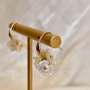 Può includere: Orecchini a cerchio color oro adornati con grappoli di delicati fiori bianco sporco. Ogni fiore presenta un centro traslucido e una piccola perla color oro. Gli orecchini sono esposti su un supporto color oro, evidenziando il loro design elegante.