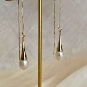 Op de afbeelding: Een paar gouden oorhangers met een witte parel aan elke oorbel. De oorhangers hangen aan een gouden standaard.