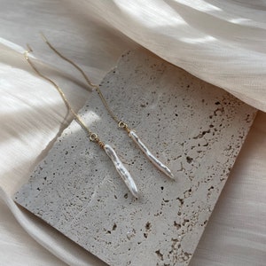 Könnte beinhalten: Ein Paar lange, zarte goldene Kettenohrringe mit weißen Perlenanhängern. Die Ohrringe werden auf einer hellbeigen, strukturierten Oberfläche präsentiert.