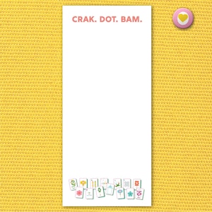 May include: A white notepad with the text "CRAK. DOT. BAM." printed in pink at the top. The notepad has a border of colorful Chinese dominoes at the bottom. A pink button with a white heart is in the top right corner.