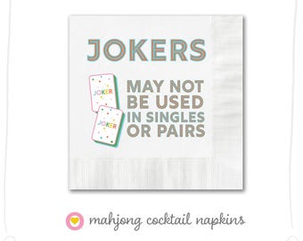 May include: White cocktail napkin with the text "JOKERS MAY NOT BE USED IN SINGLES OR PAIRS" and two images of playing cards with the word "JOKER" on them. The text is in brown and blue, and the cards are in pink, blue, green, and yellow.