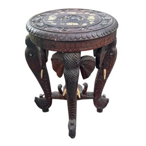 May include: A round dark brown wooden stool featuring three carved elephant legs with ivory-colored tusks. The circular top displays intricate floral carvings and inlaid light-colored decorative accents surrounding a central elephant motif.