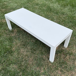 May include: A white rectangular bench with four square legs. The bench is made of a solid material and is set on a grassy surface. The bench measures approximately 60 inches long.