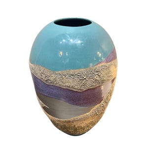 May include: A ceramic vase with a rounded shape featuring a smooth turquoise upper section, a central band of purple, and a dark grey base. The surface is accented with textured, sandy-coloured bands that create a layered, multi-dimensional effect.