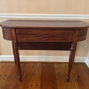 May include: A mahogany-colored, antique table with a rounded front edge. The table has a single drawer and four slender, turned legs. The table is approximately 30 inches long, 15 inches deep, and 30 inches tall.