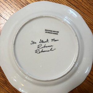 May include: A white ceramic plate with a scalloped edge. The text "The Stunt Men Rebecca Rebouchi" is handwritten in black ink on the plate's surface. The plate is marked "Dishwasher & Microwave Safe Made in China/Fabriqu&eacute; en Chine."