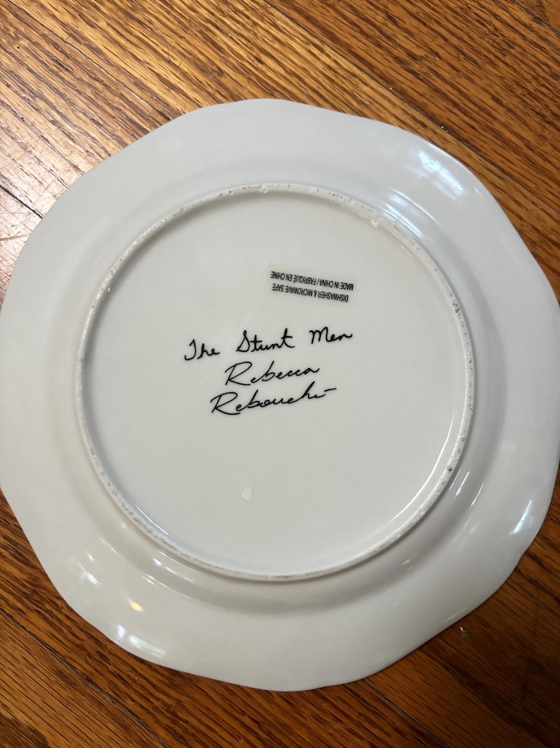 May include: A white ceramic plate with a scalloped edge. The text "The Stunt Men Rebecca Rebouchi" is handwritten in black ink on the plate's surface. The plate is marked "Dishwasher & Microwave Safe Made in China/Fabriqu&eacute; en Chine."