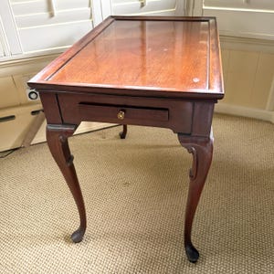May include: A mahogany-colored side table with a rectangular top and a single drawer. The table has curved legs and a brass knob on the drawer. The table measures approximately 30 inches long, 18 inches wide, and 28 inches high.