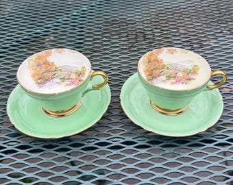 May include: Two matching vintage teacups and saucers. The cups and saucers are a light green color with gold trim. The inside of the cups feature a landscape painting of a bridge and trees. A classic tea set.