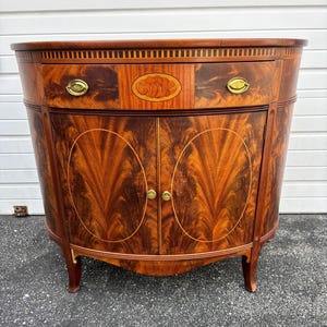 May include: A mahogany-toned cabinet with a curved front, featuring a drawer with brass hardware and an oval inlay. The cabinet has two doors with a striking wood grain pattern and brass knobs, resting on four legs.