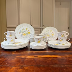 Puede incluir: Un juego de vajilla completo con temática floral que incluye platos, platillos y tazas blancos. Los platos y las tazas están adornados con un estampado floral amarillo, azul y verde. El juego incluye platos llanos, platos de postre y tazas de té.