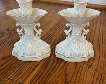May include: A pair of white porcelain candle holders with gold trim. The holders feature a scalloped design and intricate floral details.