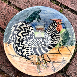 May include: A hand-painted ceramic plate featuring a rooster with black and white patterned feathers, a red comb, and yellow feet. The background depicts a landscape with a building and trees, with a blue and green sky.