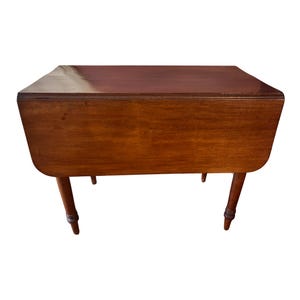 May include: A rectangular, wooden drop-leaf table with a rich, brown finish. The table has four slender, turned legs and a hinged tabletop that can be folded down. The table measures approximately 30 inches long and 20 inches wide when open.