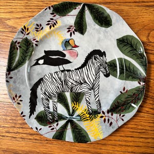 May include: A decorative ceramic plate with a zebra and a bird design. The plate features a gray background with green leaves, yellow accents, and red branches. The zebra has black and white stripes.