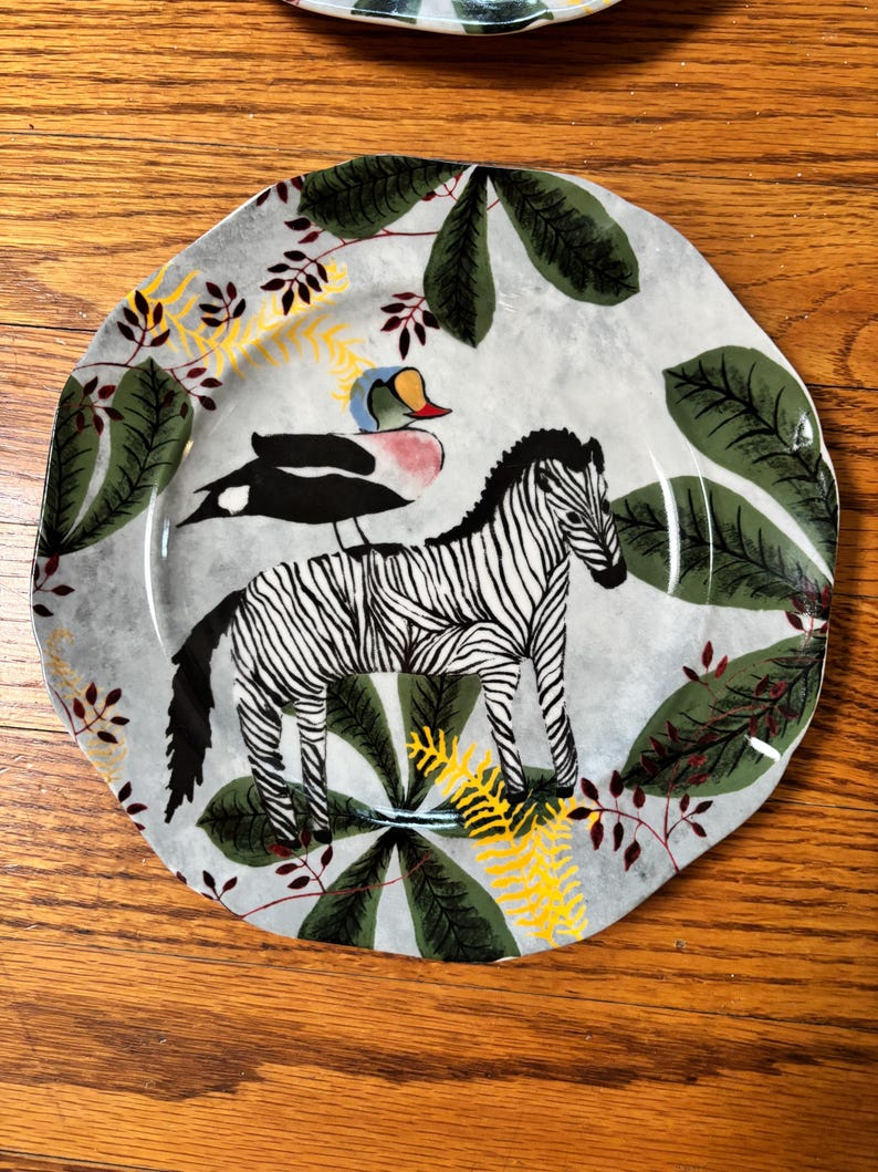 May include: A decorative ceramic plate with a zebra and a bird design. The plate features a gray background with green leaves, yellow accents, and red branches. The zebra has black and white stripes.