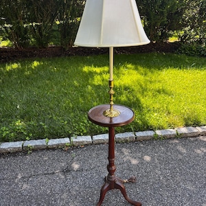 May include: A vintage floor lamp with a cream-colored lampshade and a dark brown wooden base. The lamp features a small round table and ornate brass accents. The lamp stands approximately 60 inches tall.