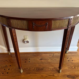 May include: A half-moon shaped wooden console table with a dark brown finish. The table features a decorative gold-colored panel and a small drawer with a detailed handle. The legs are slender and have gold-colored accents at the feet.