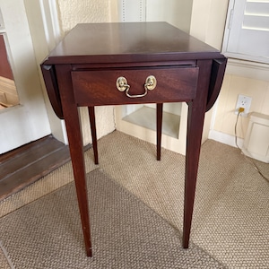 May include: A dark cherry-finished drop-leaf table with a single drawer and brass hardware. The table has four slender legs and is approximately 30 inches tall. The tabletop is rectangular with rounded corners and drop leaves on each side.