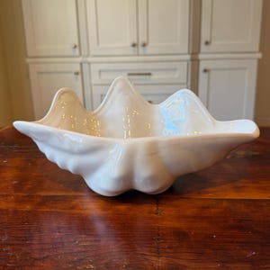 May include: A large, white ceramic clam shell-shaped bowl. The bowl has a glossy finish and a scalloped edge, with a textured surface. The bowl is sitting on a dark wooden surface.