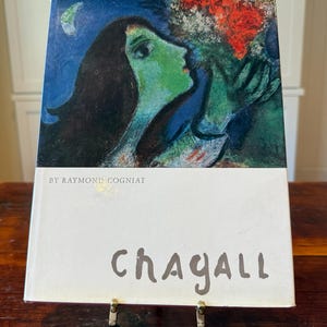 May include: A book titled "Chagall" by Raymond Cogniat, displayed on a stand. The cover features a colorful painting of two figures with green and blue tones. The word "Chagall" is written in a handwritten style.