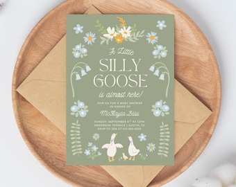 Pode incluir: Um convite para chá de bebé com fundo verde e o texto "A Little Silly Goose is almost here!". O convite apresenta ilustrações florais e de gansos, e é colocado num prato de madeira com um envelope castanho.