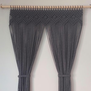 May include: A grey macrame curtain set, suspended from a natural wood rod. The top features a decorative woven pattern, with long, vertical strands forming the curtain panels. The panels are gathered with woven bands. The curtain is hung against a white wall.