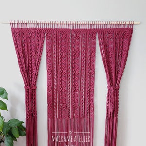 Pode incluir: Três tapeçarias de macramé cor de vinho com franjas, penduradas em uma haste de madeira. A peça central apresenta um padrão geométrico de diamantes. O texto "MACRAME ATELIER by Ines" é visível na parte inferior da peça central.