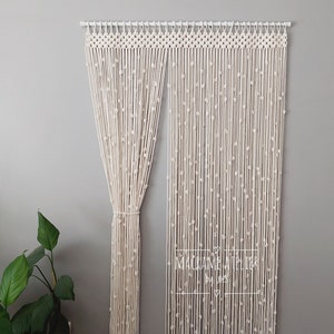 K&ouml;nnte beinhalten: Ein wei&szlig;er Makramee-Vorhang mit Perlenmuster h&auml;ngt an einer wei&szlig;en Stange. Der Vorhang ist aus Baumwollschnur gefertigt und hat einen schlichten, minimalistischen Stil. Der Text "MACRAME  ATELIER by LNES" ist in der unteren rechten Ecke des Bildes sichtbar.