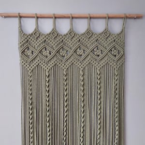 May include: A macrame wall hanging with a geometric pattern in a sage green color. The hanging is made of cotton cord and is attached to a wooden dowel rod.