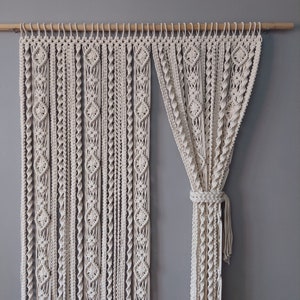 Può includere: Una tenda in macramè bianca con un motivo intrecciato è appesa a un'asta di legno. La tenda è legata indietro su un lato, creando un effetto drappeggiato.