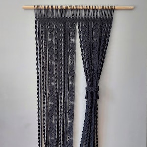 May include: Black macrame curtain with a wooden dowel rod at the top. The curtain is made of multiple strands of cord that are knotted together in a variety of patterns.