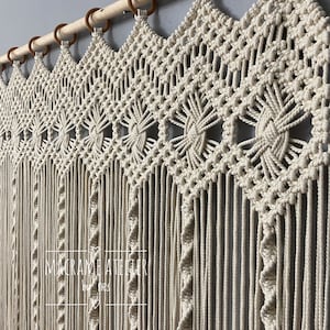 Puede incluir: Un tapiz de macramé blanco con un patrón geométrico. El tapiz está hecho de cordón de algodón y está sujeto a una varilla de madera con anillos de madera marrón. El tapiz tiene una franja en la parte inferior.