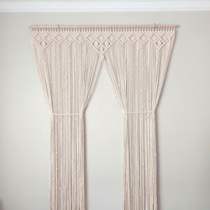 May include: Cream-coloured macrame curtain panel with a geometric design at the top. The curtain hangs from a wooden rod and features vertical strands with small, decorative knots. The panel is tied in the middle with a matching cord.