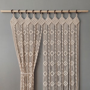 Può includere: Una tenda in macramè bianca con un'asta di legno e anelli di metallo nero. La tenda è realizzata con una serie di corde annodate che creano uno schema geometrico.