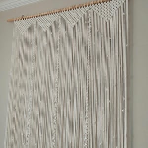 Puede incluir: Cortina de macramé color crema colgada de una barra de madera. La cortina presenta un diseño geométrico en la parte superior y hebras verticales con detalles anudados. Esta pieza decorativa añade un toque bohemio a cualquier habitación.