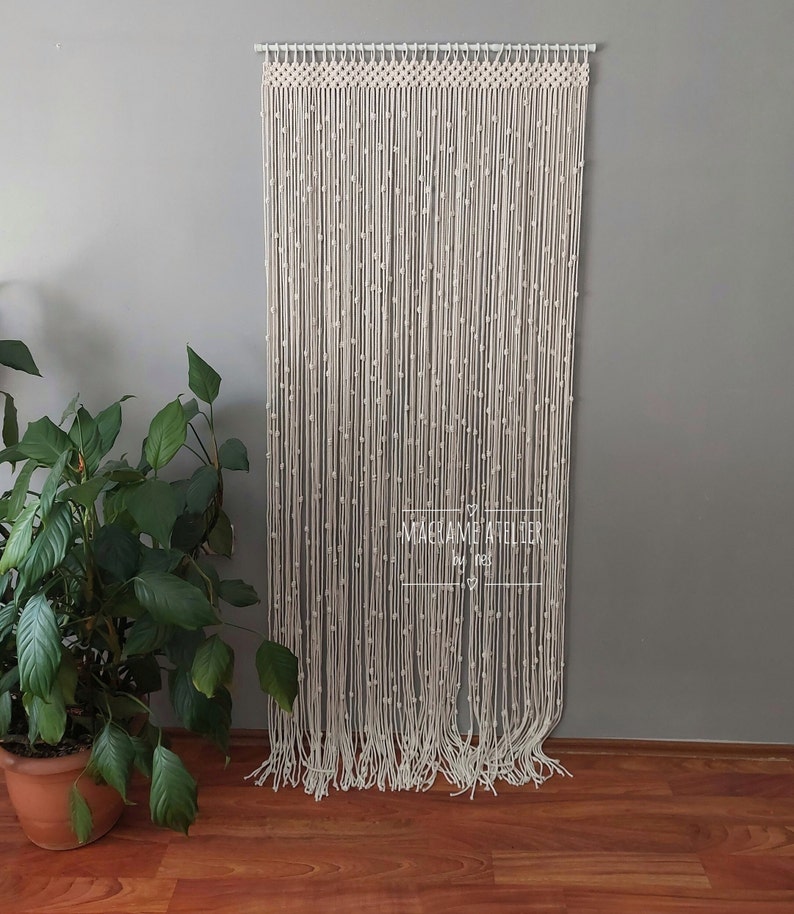 K&ouml;nnte beinhalten: Ein wei&szlig;er Makramee-Wandbehang mit einer Perlenfransen. Der Behang ist aus nat&uuml;rlichem Baumwollschnur gefertigt und etwa 178 cm hoch. Der Makramee h&auml;ngt an einer wei&szlig;en Wand mit einer Pflanze in einem Terrakotta-Topf im Vordergrund.